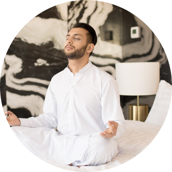 A man dressed in white sits cross-legged near a body of water, with eyes closed and meditating