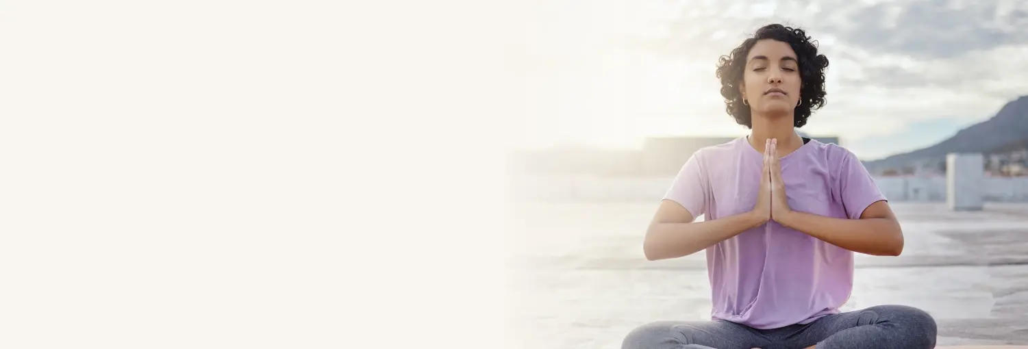 A person in a purple shirt, seated cross-legged outdoors, with eyes closed and hands in prayer position for Shakti Kriya program.