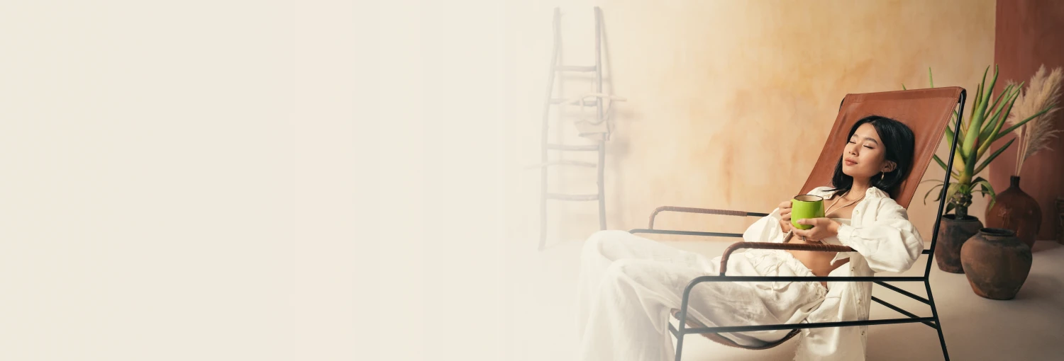 A woman sits on a couch, eyes closed, holding a mug, bathed in sunlight. Representing calm and relaxation from a deep sleep and anxiety relief course