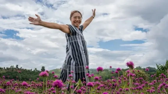 A smiling woman stands with arms outstretched in a vibrant field of pink flowers, surrounded by lush greenery and a cloudy blue sky
