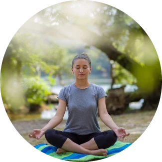 A young woman sits cross-legged on a colorful mat in a peaceful outdoor setting, eyes closed in meditation