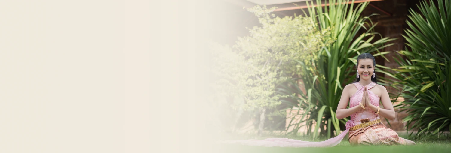 A smiling woman in a pink traditional Thai dress sitting on grass, performing the 'wai' greeting gesture.