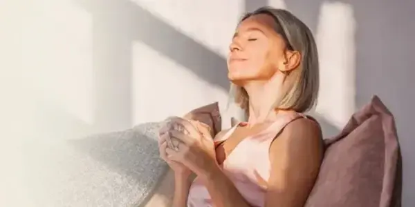 A woman sits on a couch, eyes closed, holding a mug, bathed in sunlight. Representing calm and relaxation from a deep sleep and anxiety relief course